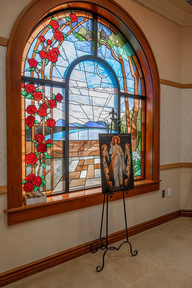 A stained-glass window featuring red roses and a landscape behind a framed portrait displayed on a metal easel.