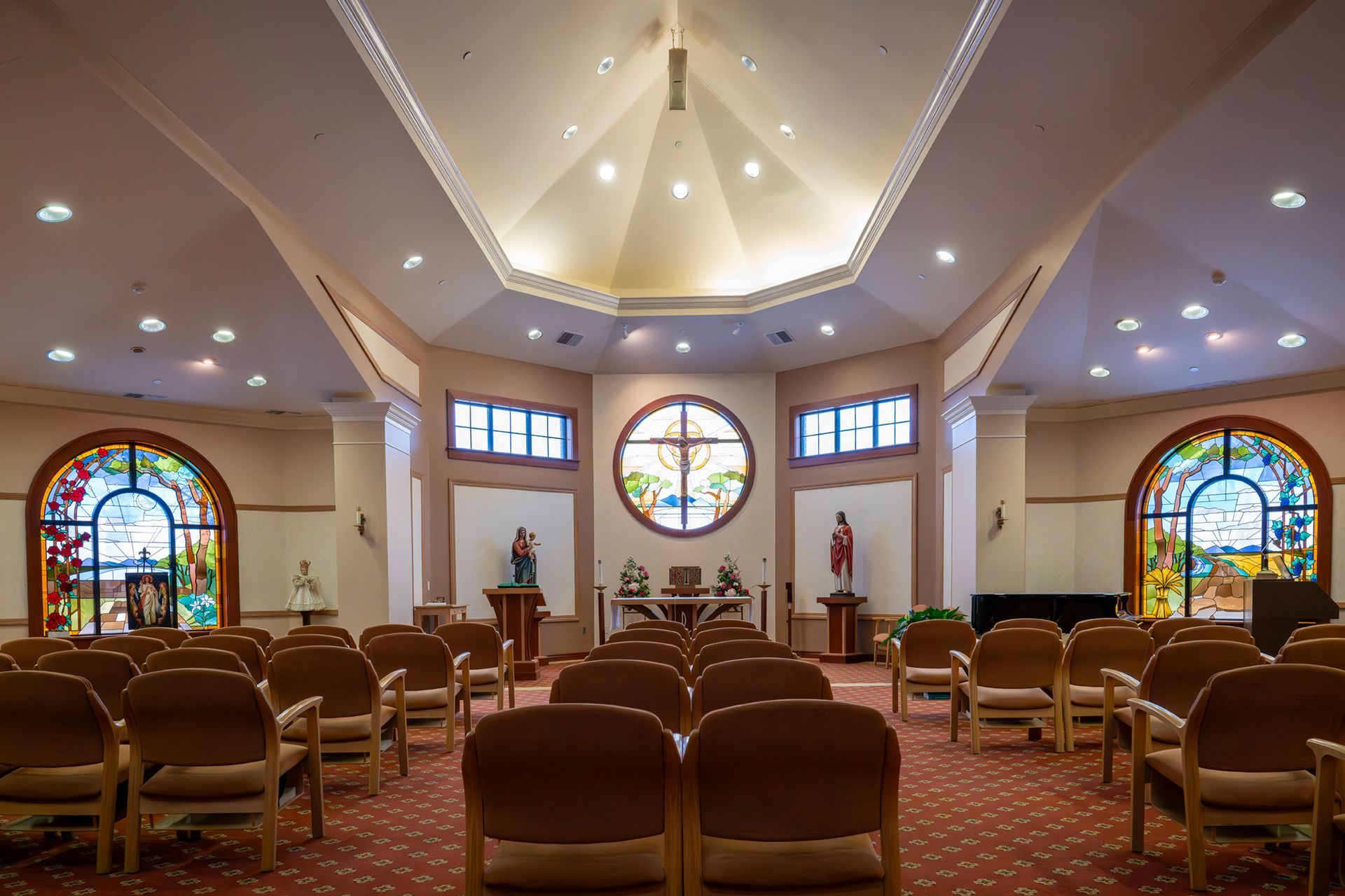 An interior view of a chapel featuring rows of chairs, a vaulted ceiling, and stained-glass windows on the walls.
