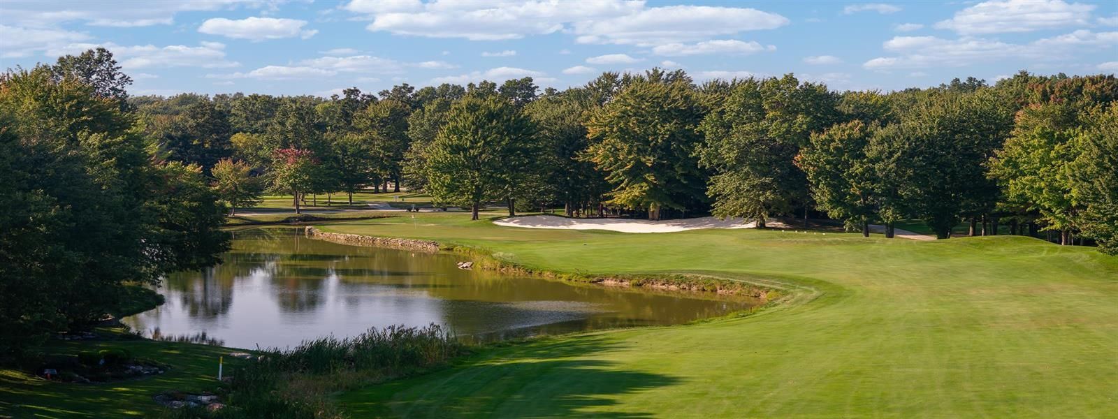 A golf course hole featuring a pond in the foreground, a grassy fairway, a sand bunker, and a dense forest background.
