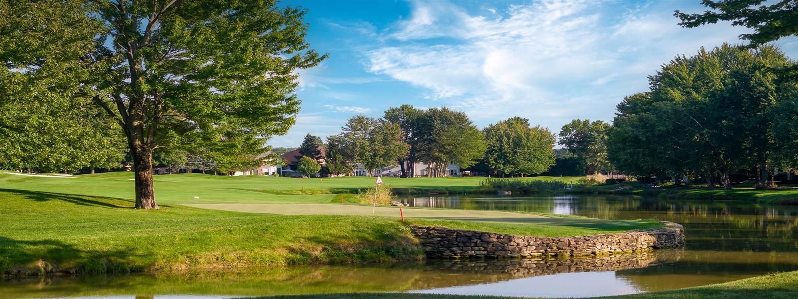 A green golf course hole features a pond in the foreground, a stone bulkhead, and lush trees under a bright blue sky.