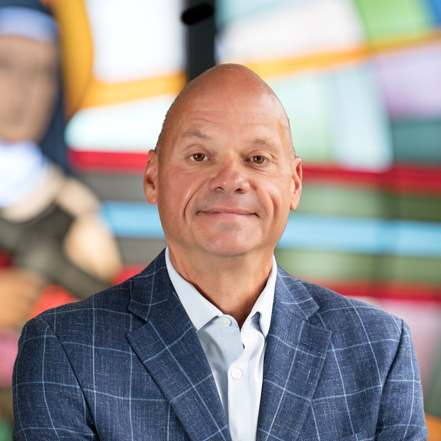 A person wearing a blue checkered blazer and a light shirt stands in front of a colorful, blurred artistic background.