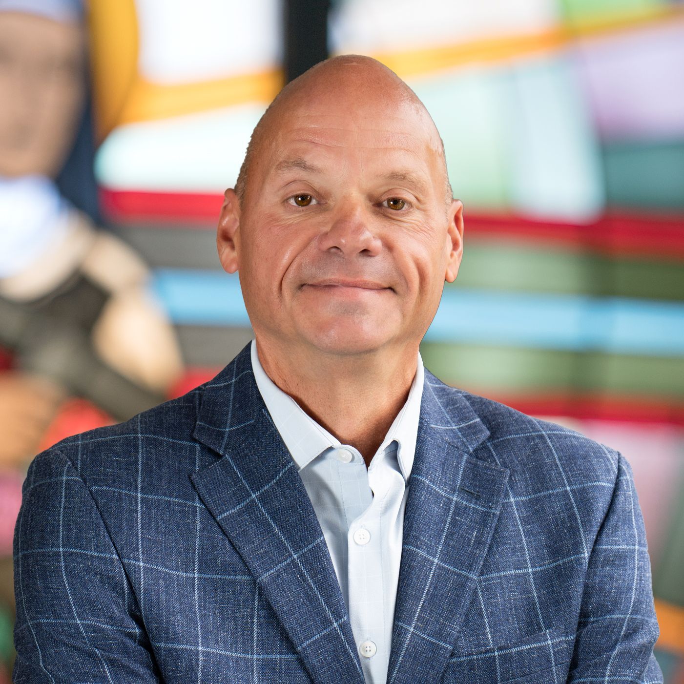 A person with a neutral expression wears a blue plaid blazer over a light-colored shirt against a colorful, blurred backdrop.