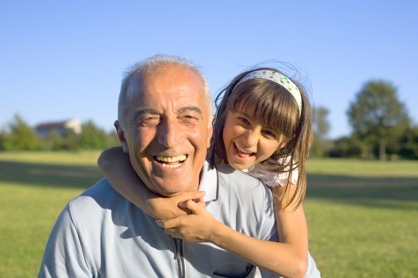 A person smiling and giving a piggyback ride to a child in a sunny, green park.