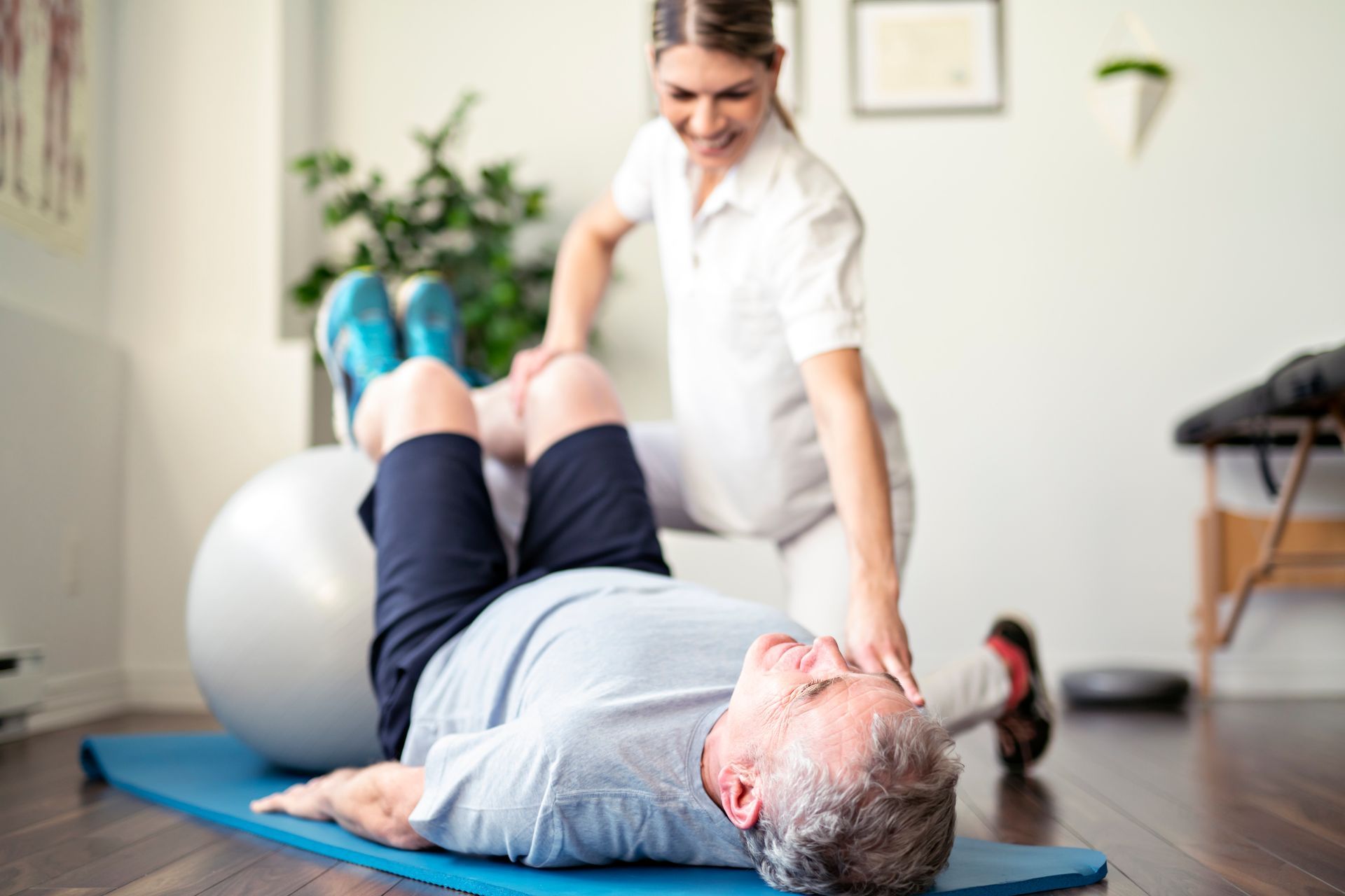 A physical therapist assists a person lying on a blue exercise mat with leg exercises using a large grey stability ball.