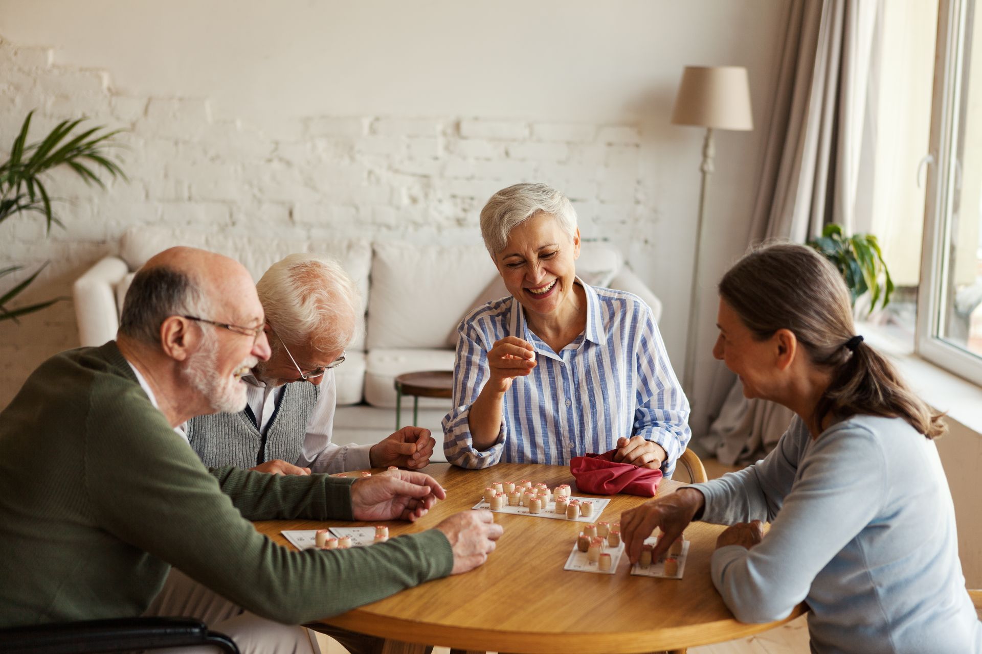 Four people sit around a wooden table in a bright room, laughing while playing a game with small tokens.