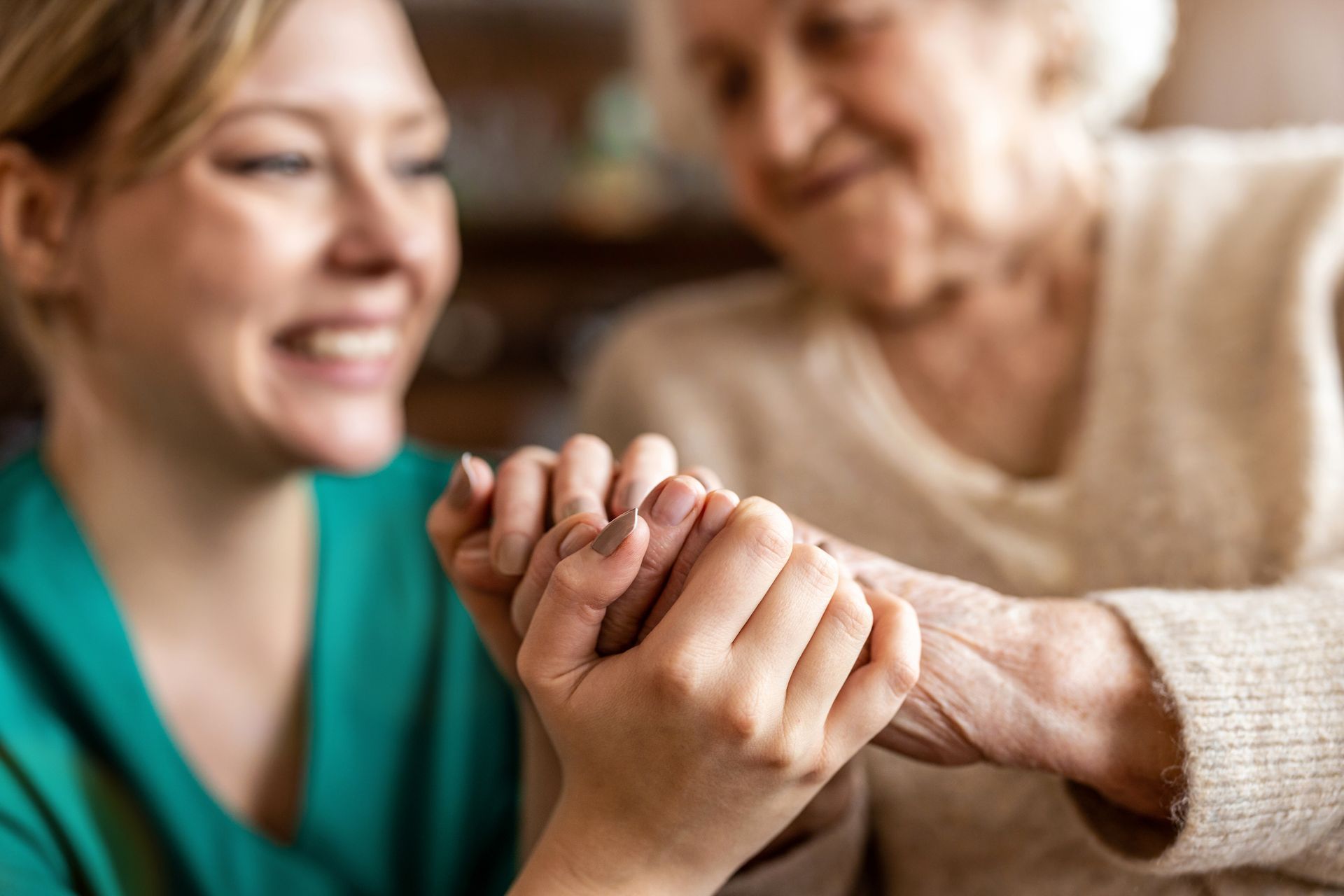 A person in green scrubs gently holds the hands of an older person, both smiling softly in an indoor setting.