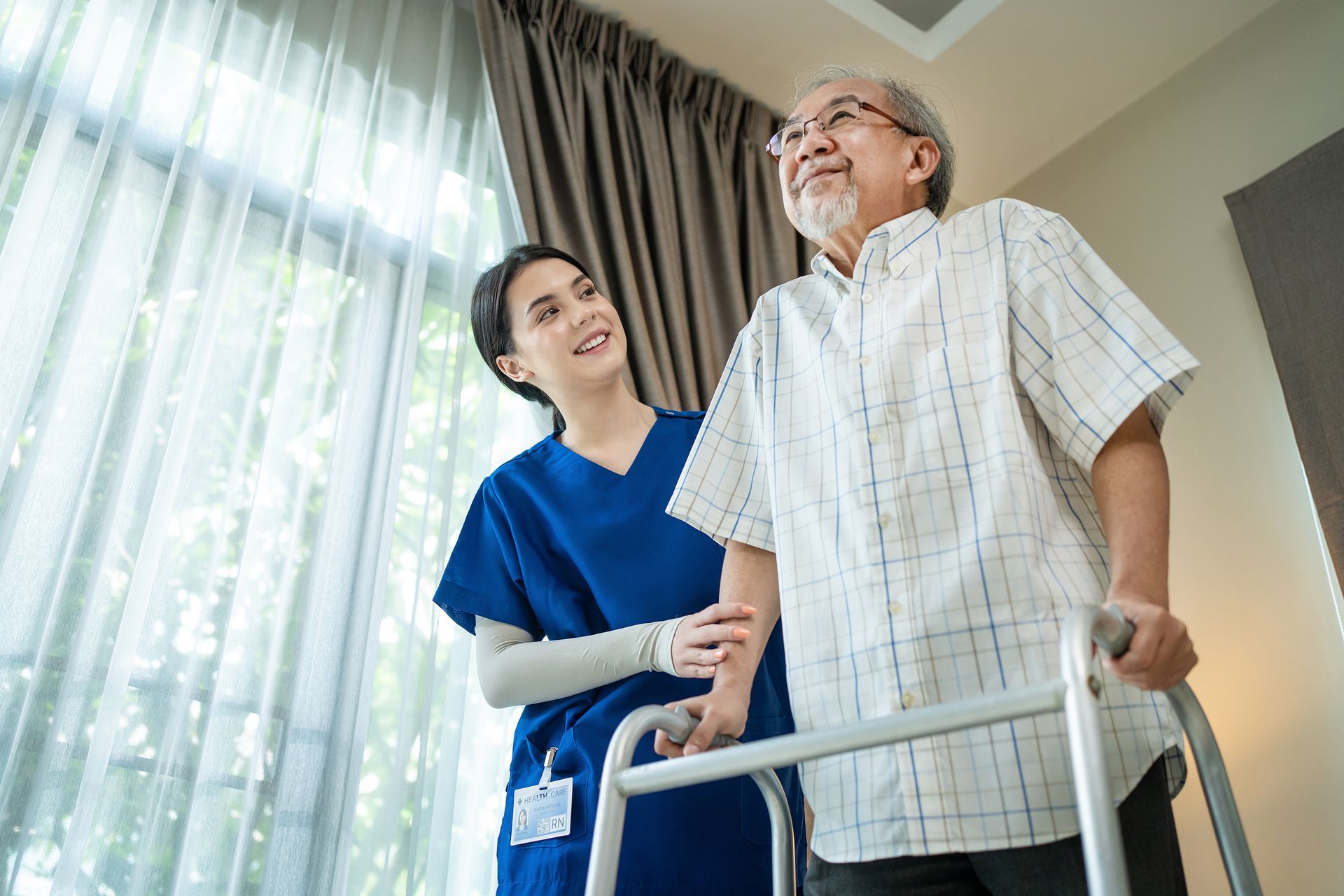A smiling healthcare worker in blue scrubs supports a man using a walker in a room with curtains.