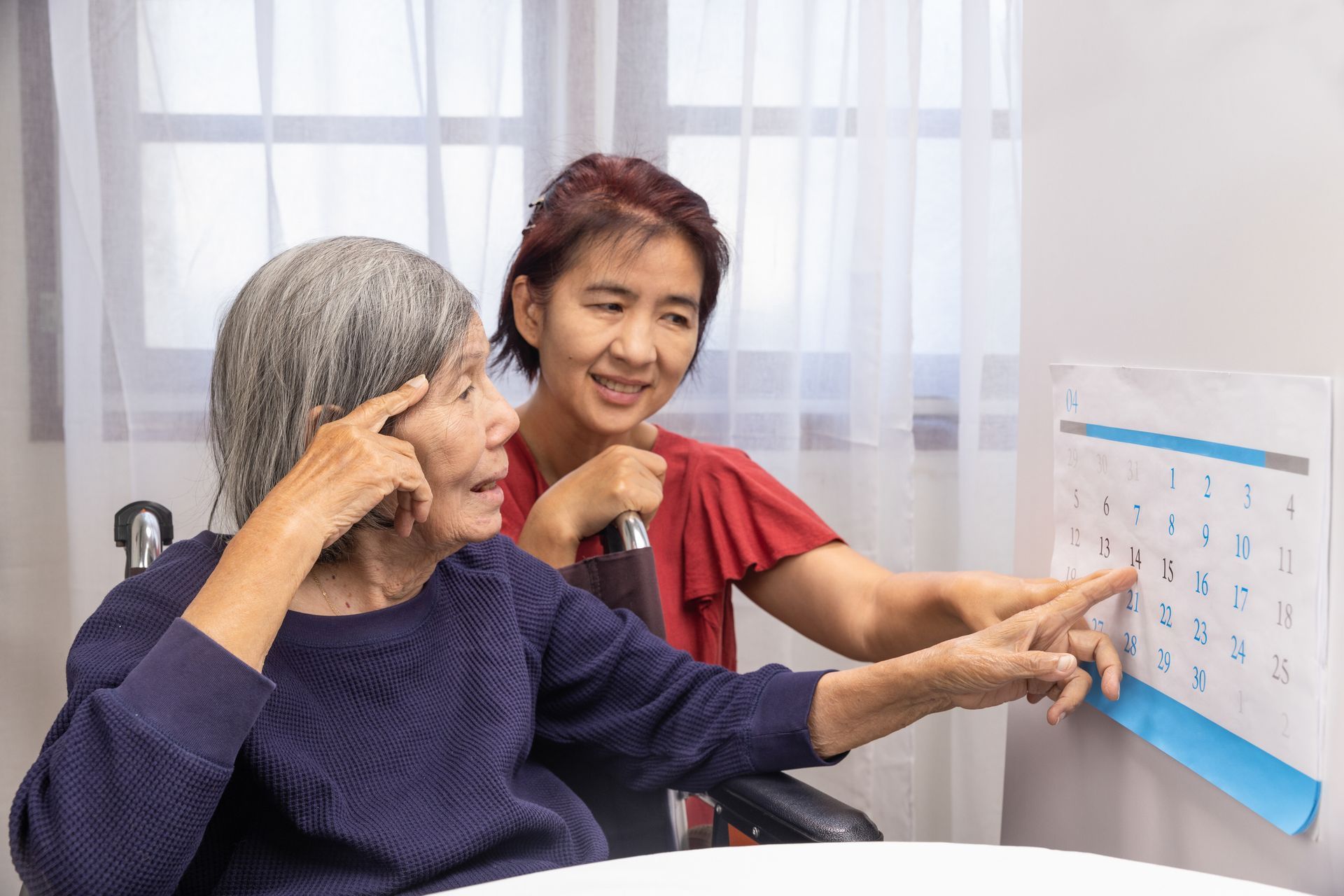 A person points to a date on a wall calendar while another person in a wheelchair watches and touches their head.