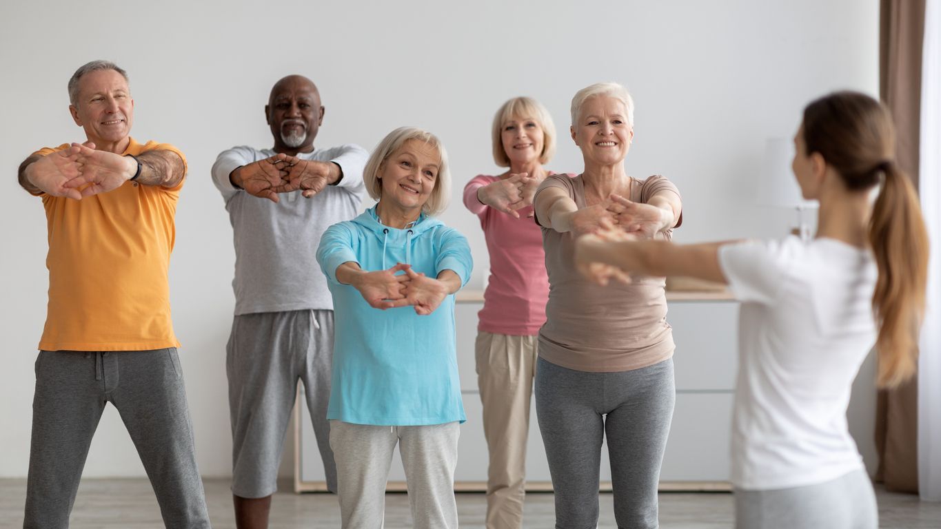 An instructor leads a group of people in a light stretching exercise in a brightly lit room.