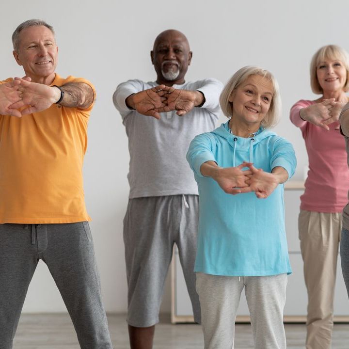 Four people standing in a row, smiling while performing an arm-stretching exercise in a light-filled room.