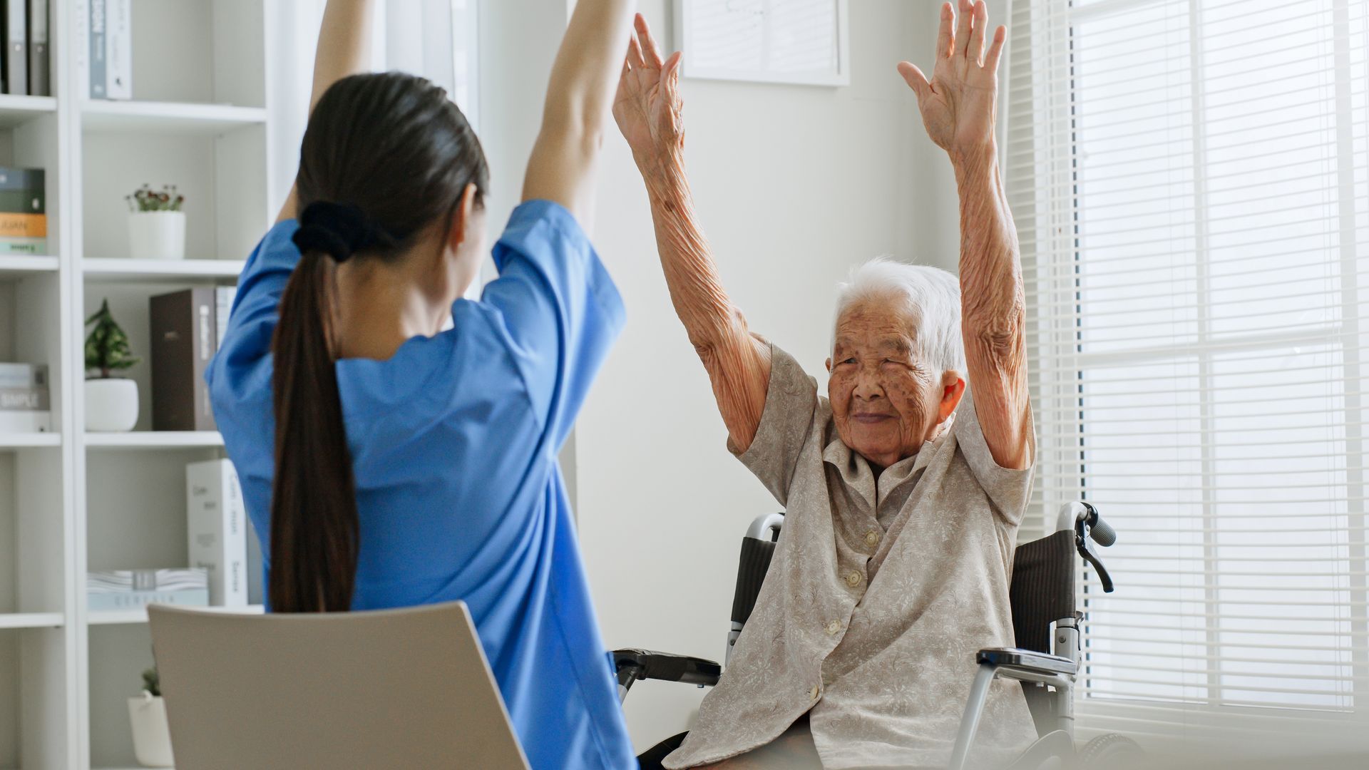 A caregiver in blue scrubs and a person in a wheelchair sit facing each other, both raising their arms for physical therapy.
