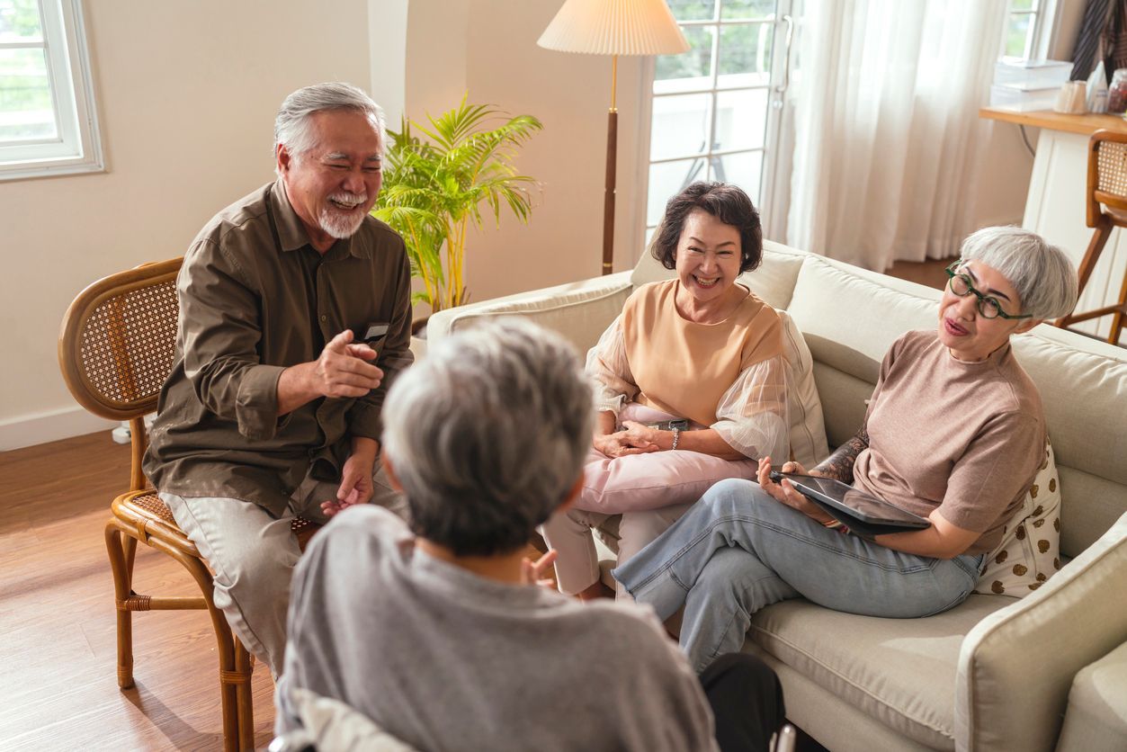 Four people sit in a living room, smiling and talking while seated on a sofa and wooden chairs.