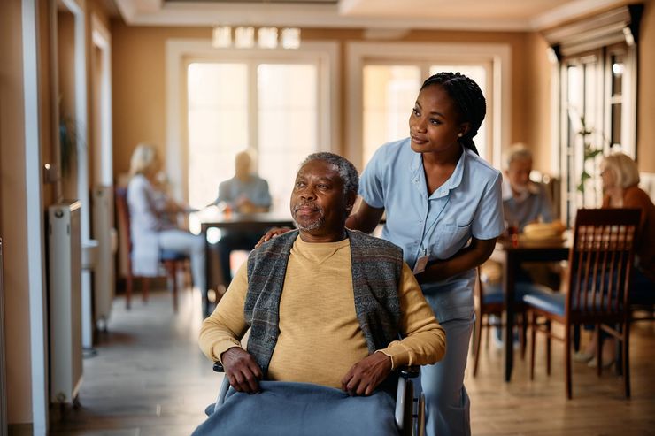 A caregiver in blue scrubs stands behind an individual in a wheelchair, moving through a brightly lit common room.