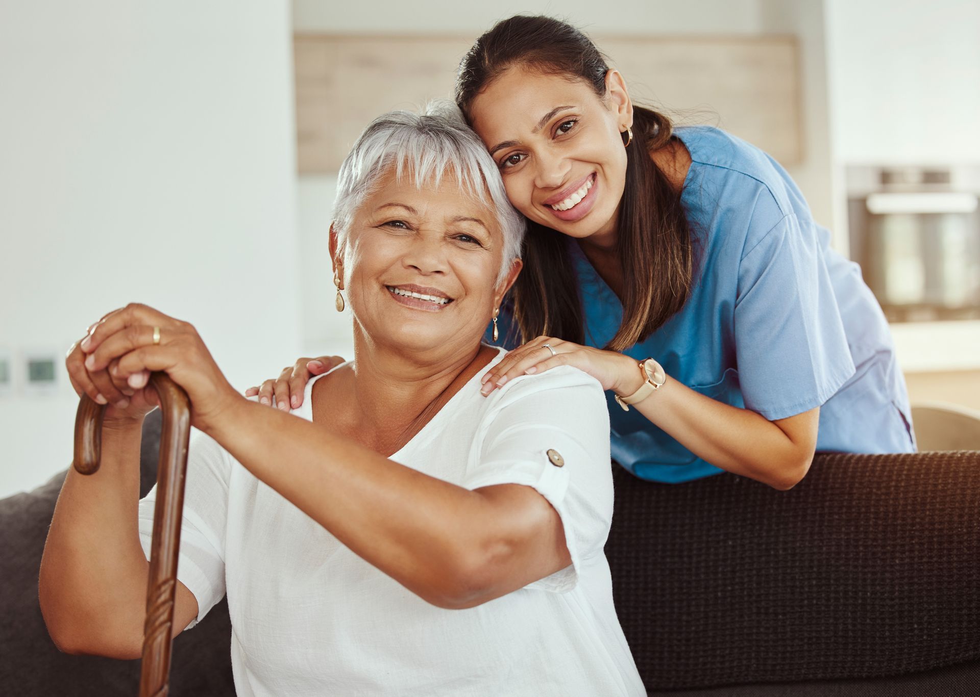 A smiling caregiver in a blue scrub top leans over a person holding a wooden cane, both looking toward the camera.