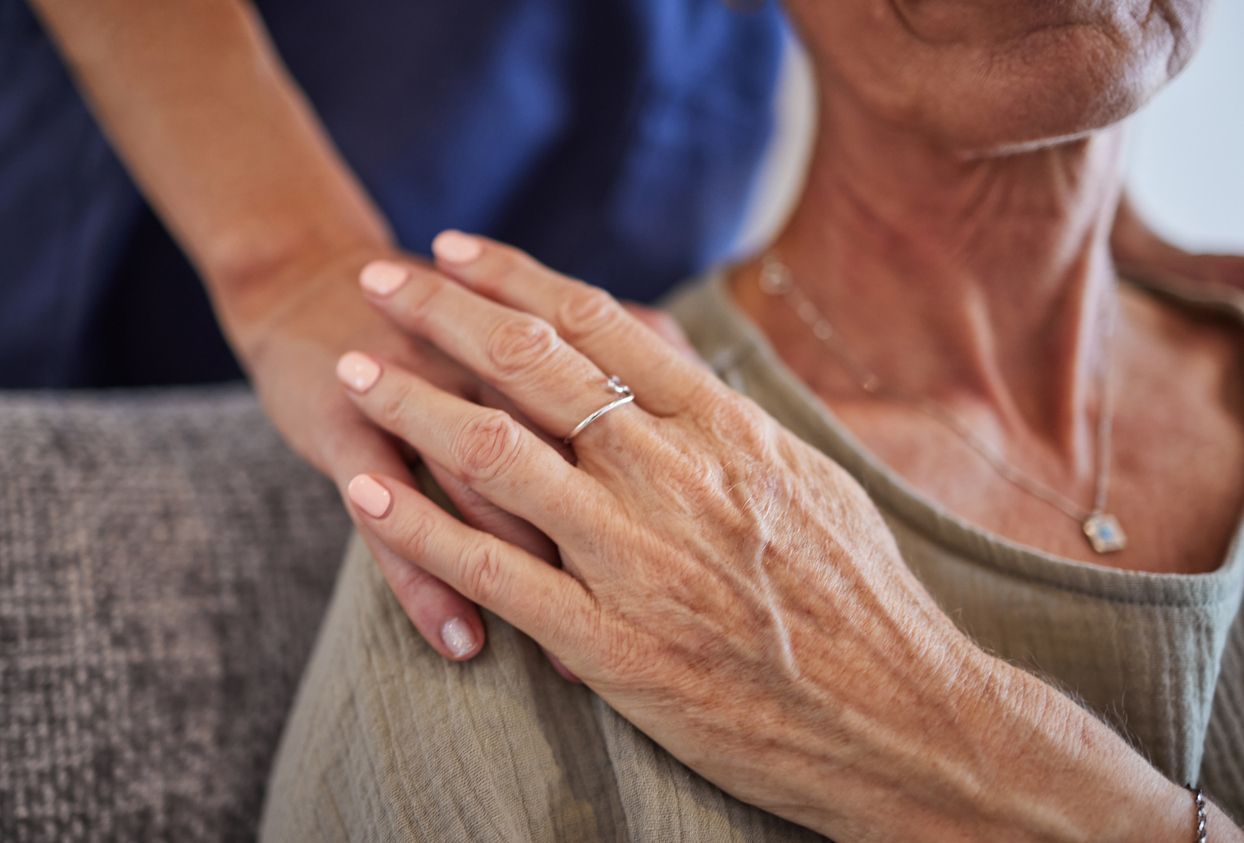 A person’s hand rests gently on the shoulder of another person wearing a necklace and a textured top.