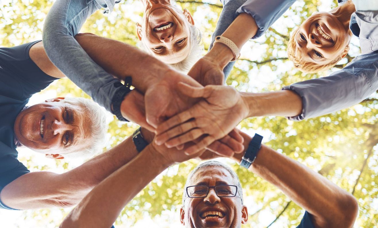 Four people standing in a circle, looking down while stacking their hands together in a show of unity outdoors.