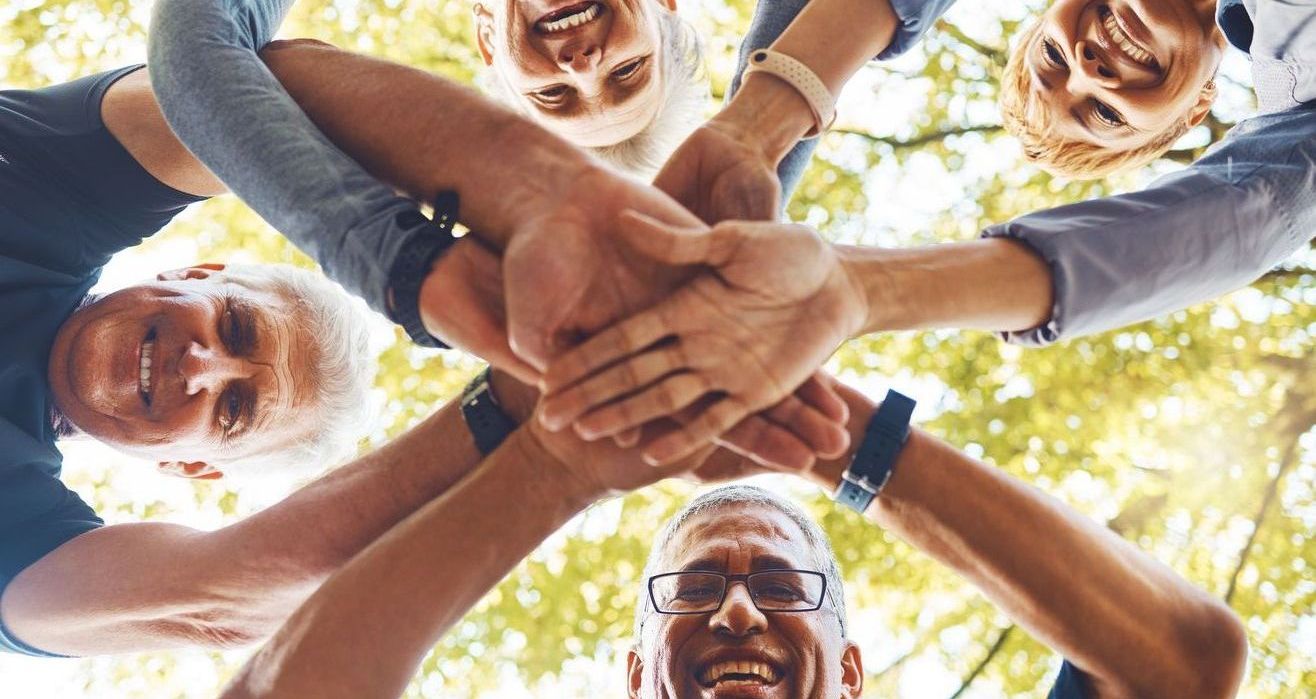 A group of people standing in a circle with their hands stacked together in the center against a bright, leafy background.