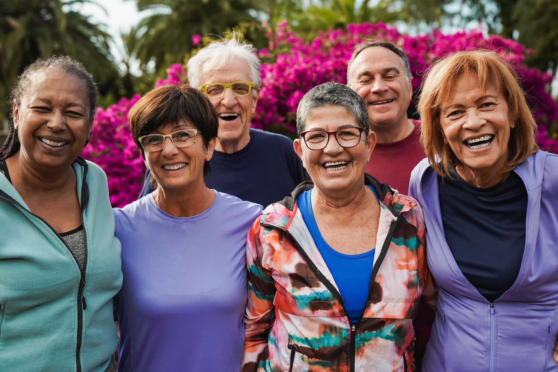 A diverse group of six smiling people stand together outdoors in front of vibrant pink flowering bushes.