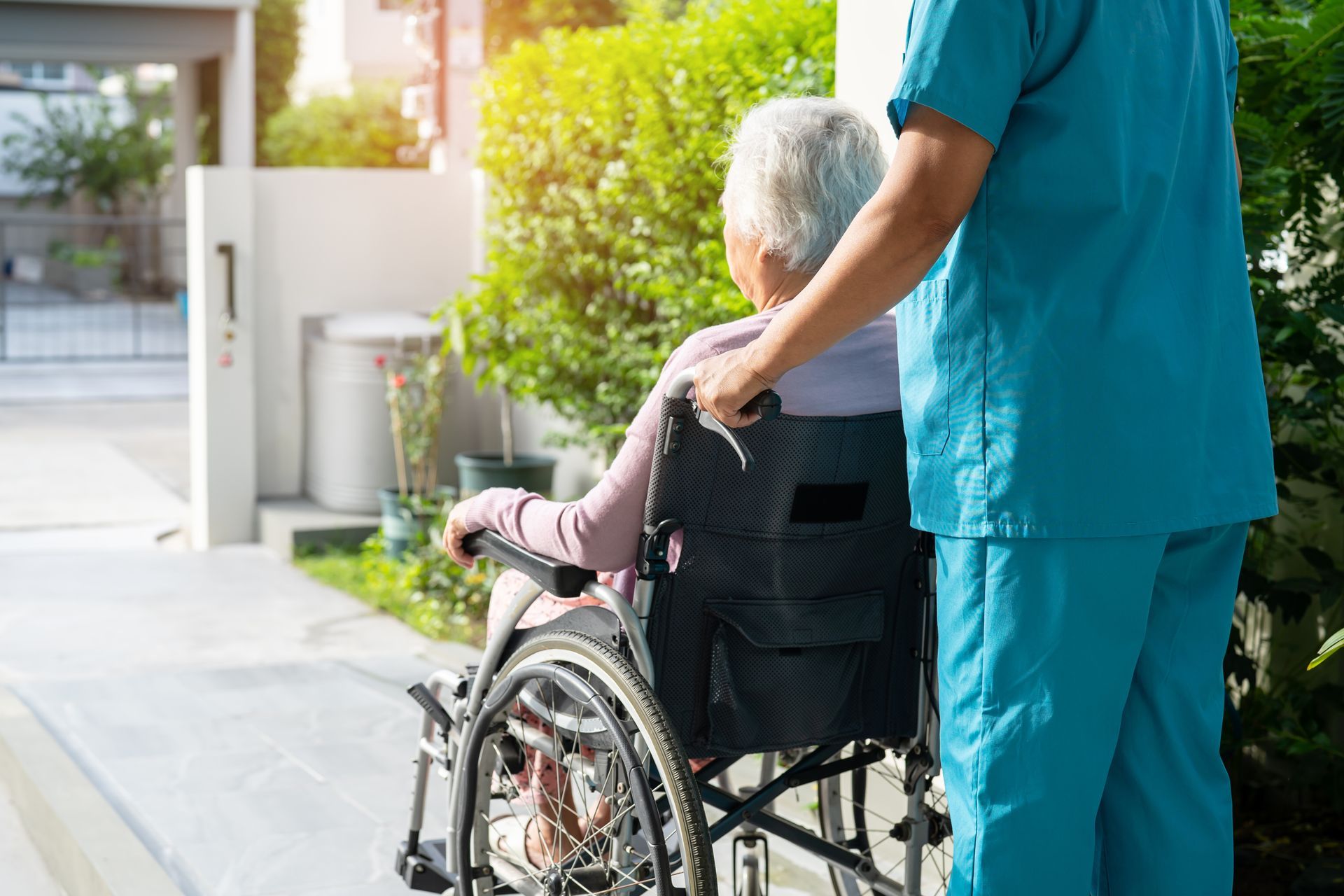 A person in a blue uniform pushes an individual in a wheelchair down an outdoor walkway.