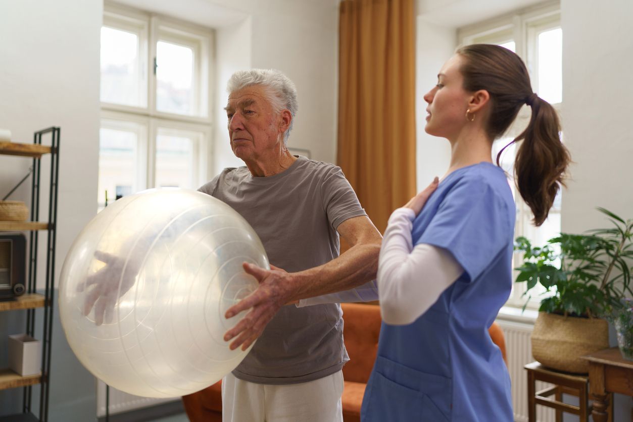 A physical therapist guides an older man through a therapeutic exercise using a large, clear inflatable exercise ball.
