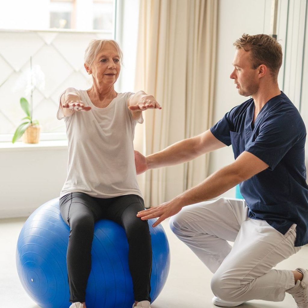 A physical therapist assists an individual with arm extension exercises while they sit on a blue exercise ball.
