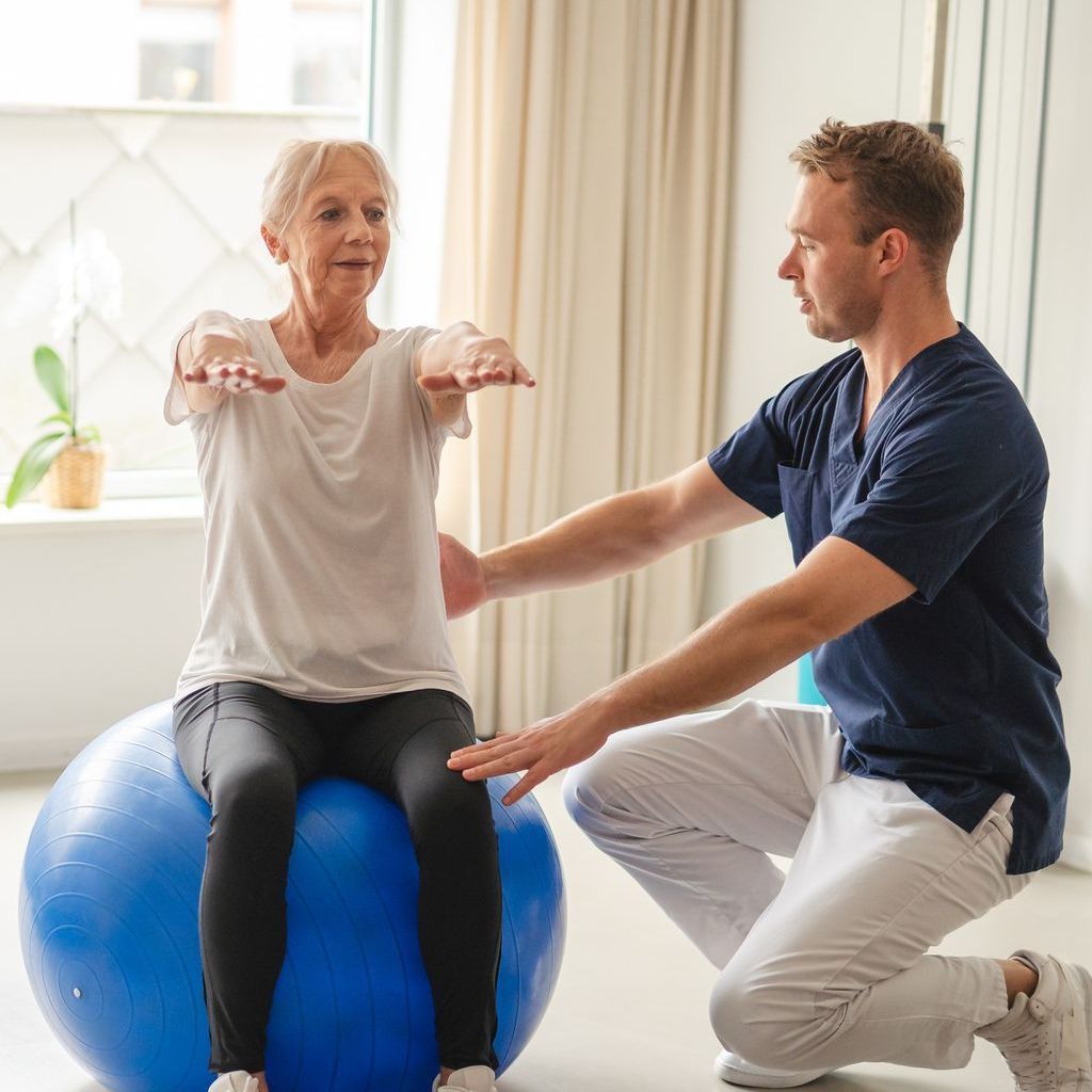 A physical therapist kneels beside a patient seated on a blue exercise ball, guiding them through an arm-raising exercise.