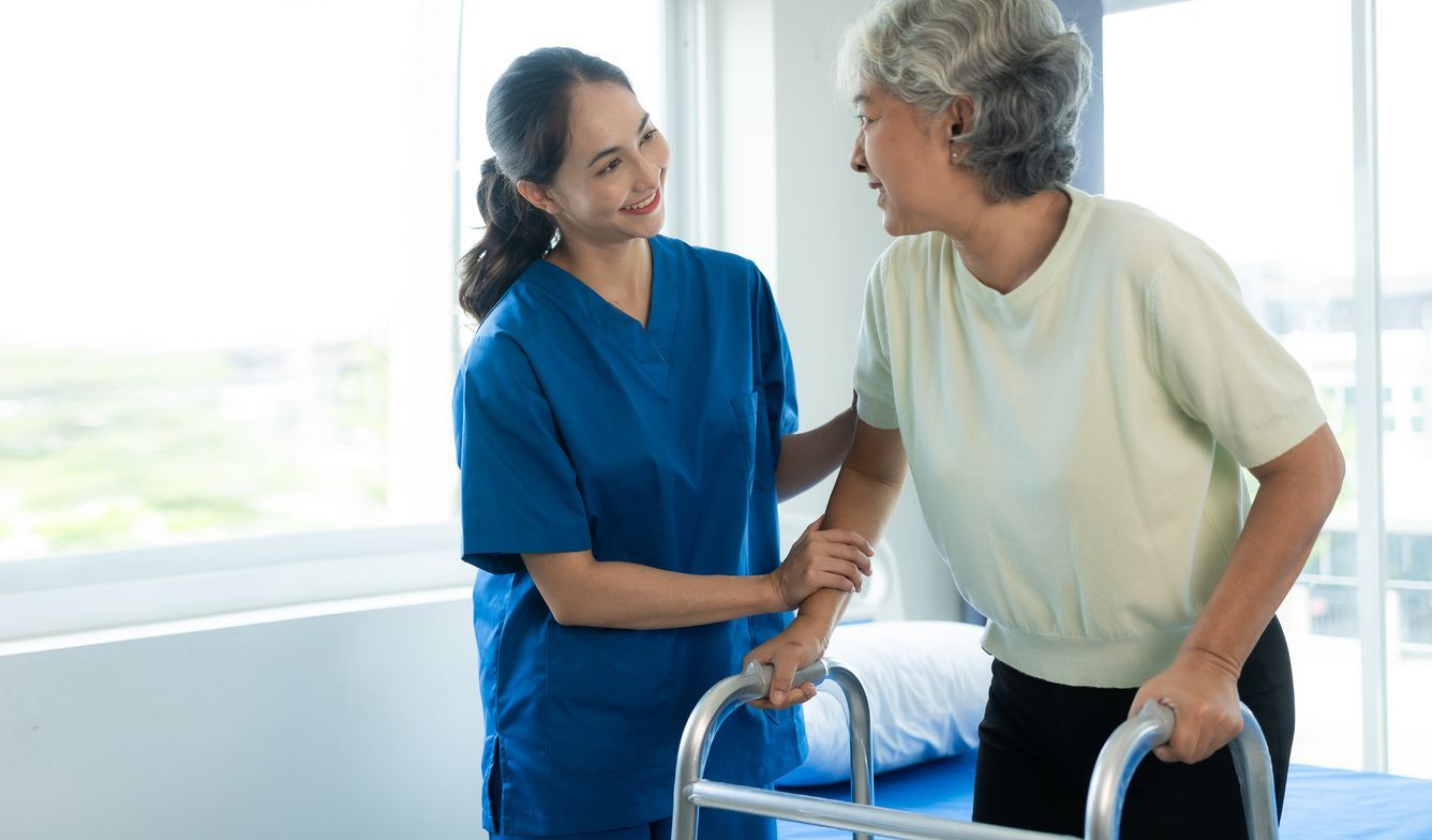 A healthcare worker in blue scrubs supports a person using a walker in a bright room with a bed.