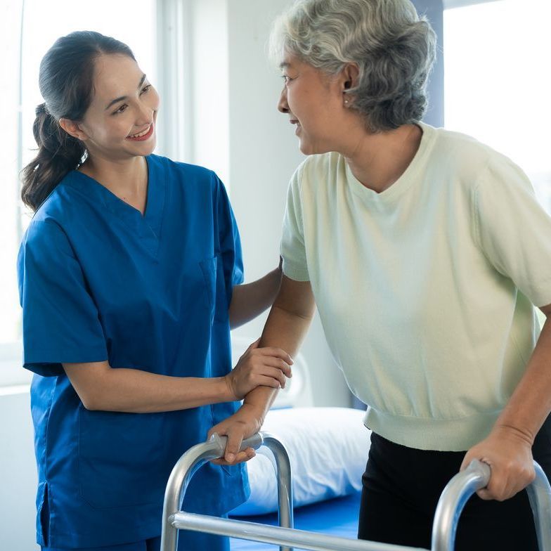 A healthcare worker in blue scrubs supports an individual using a walker in a room.