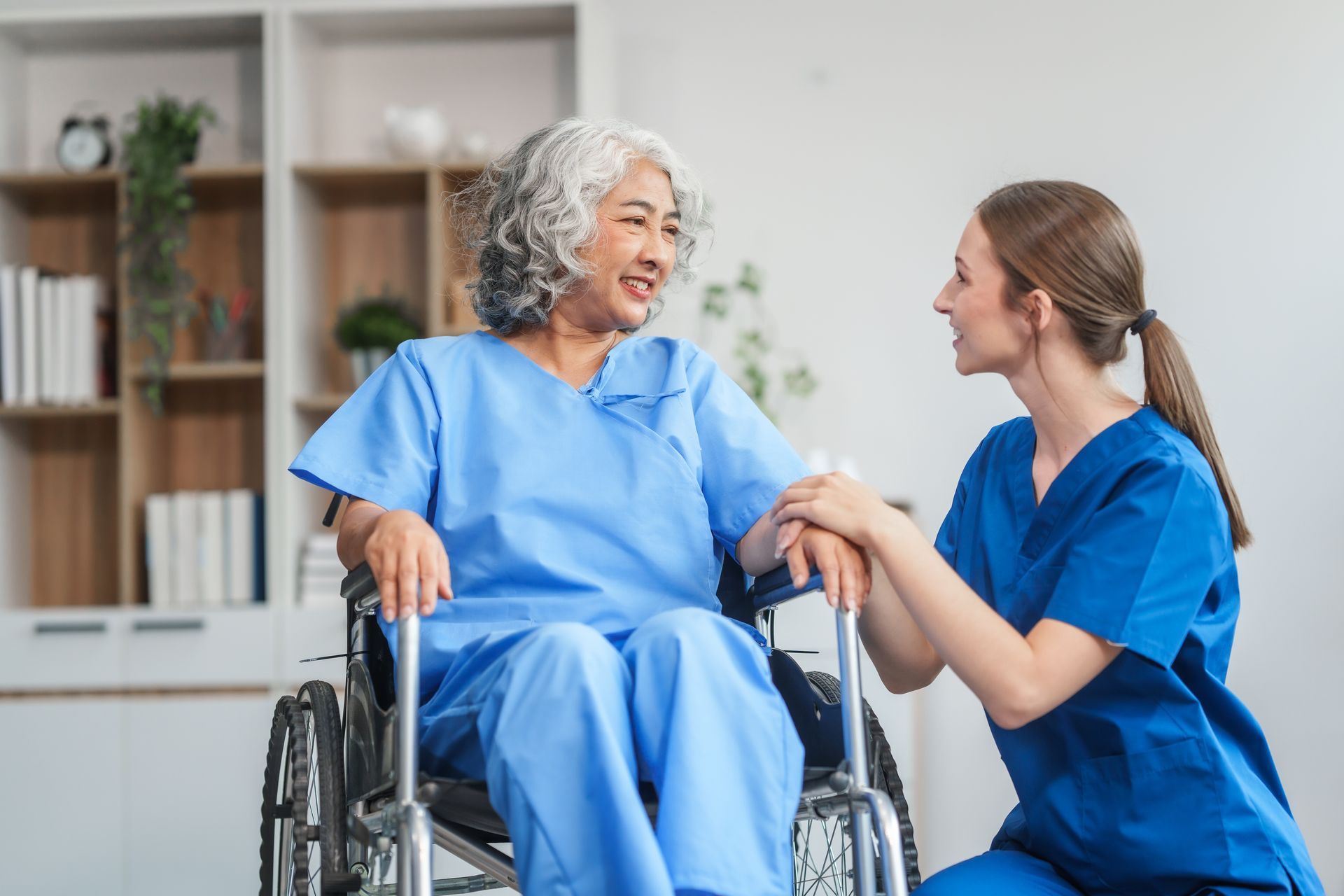 A smiling person in a blue hospital gown sits in a wheelchair while a person in blue scrubs holds their hands.