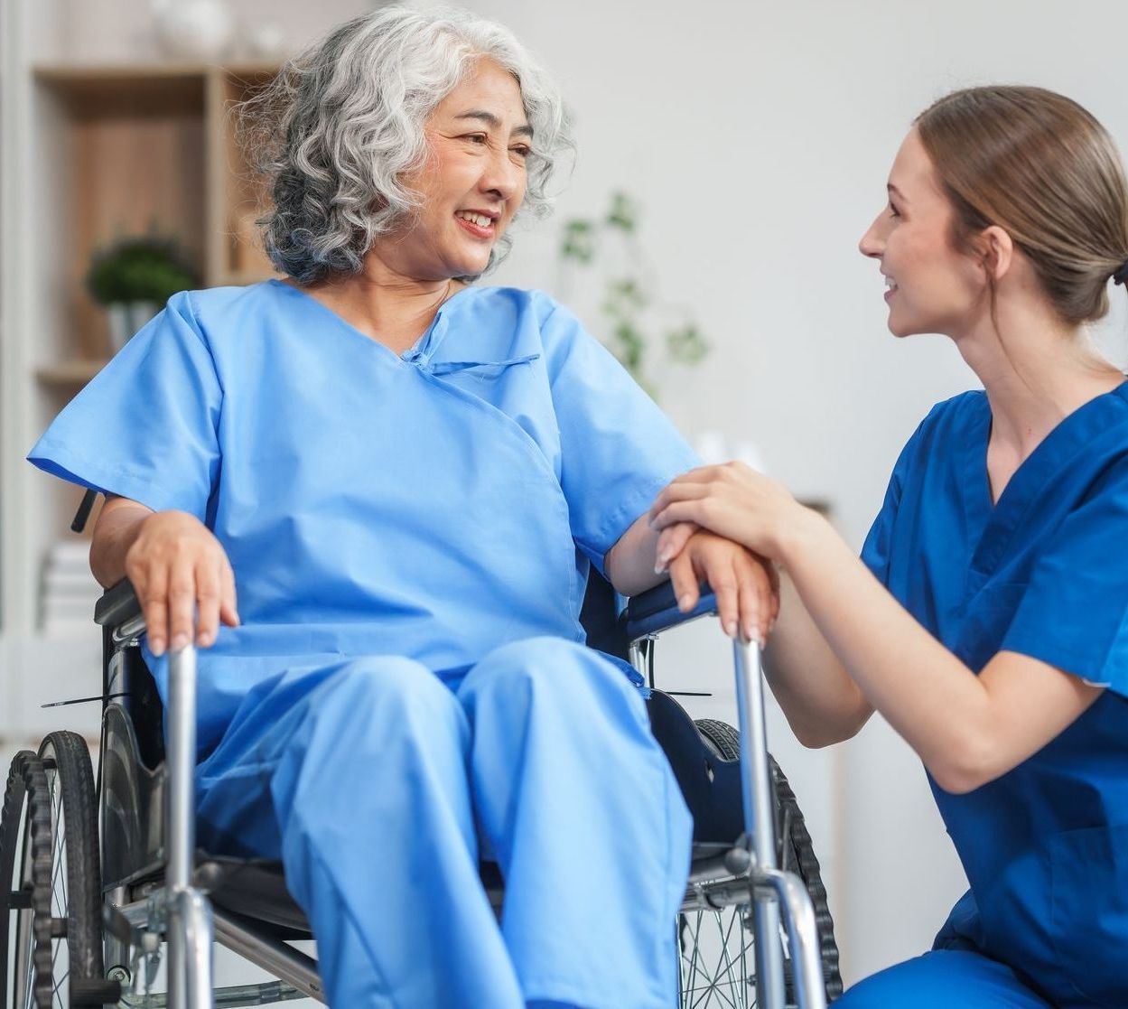 A healthcare worker in blue scrubs holds the hand of a patient in a wheelchair while smiling and speaking together.