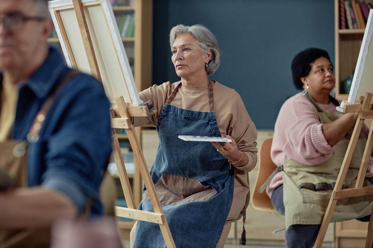 Three people in aprons stand at easels in a classroom, painting on canvases.