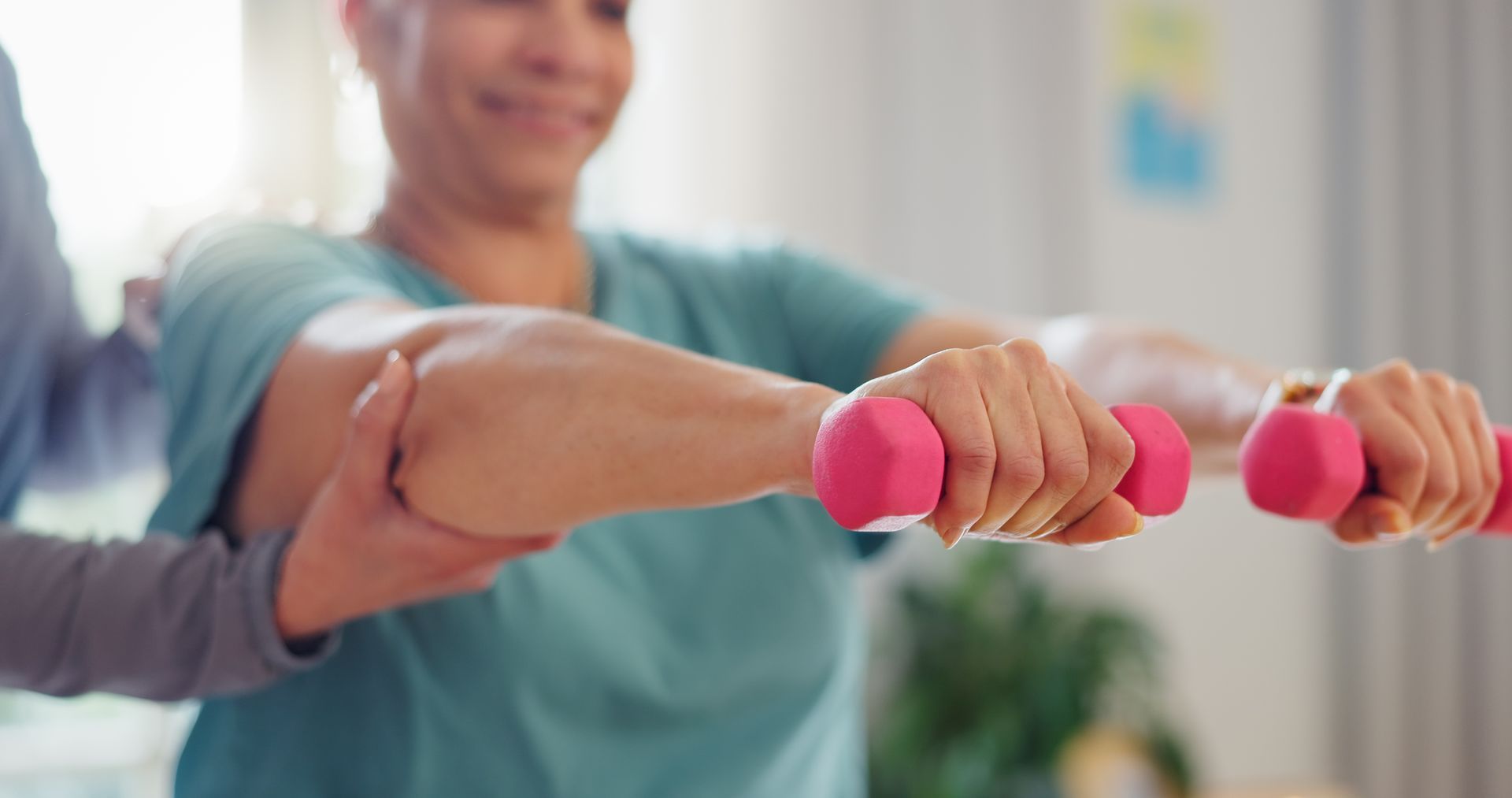 A caregiver supports a person’s arm while they hold pink dumbbells to exercise in a home setting.