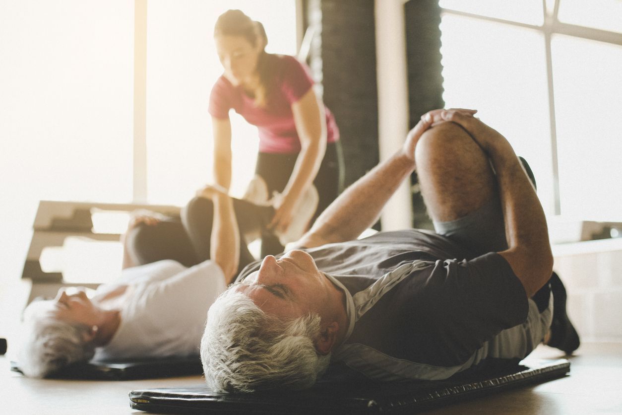 A professional instructor assists two people lying on exercise mats, stretching their legs toward their chests in a studio.
