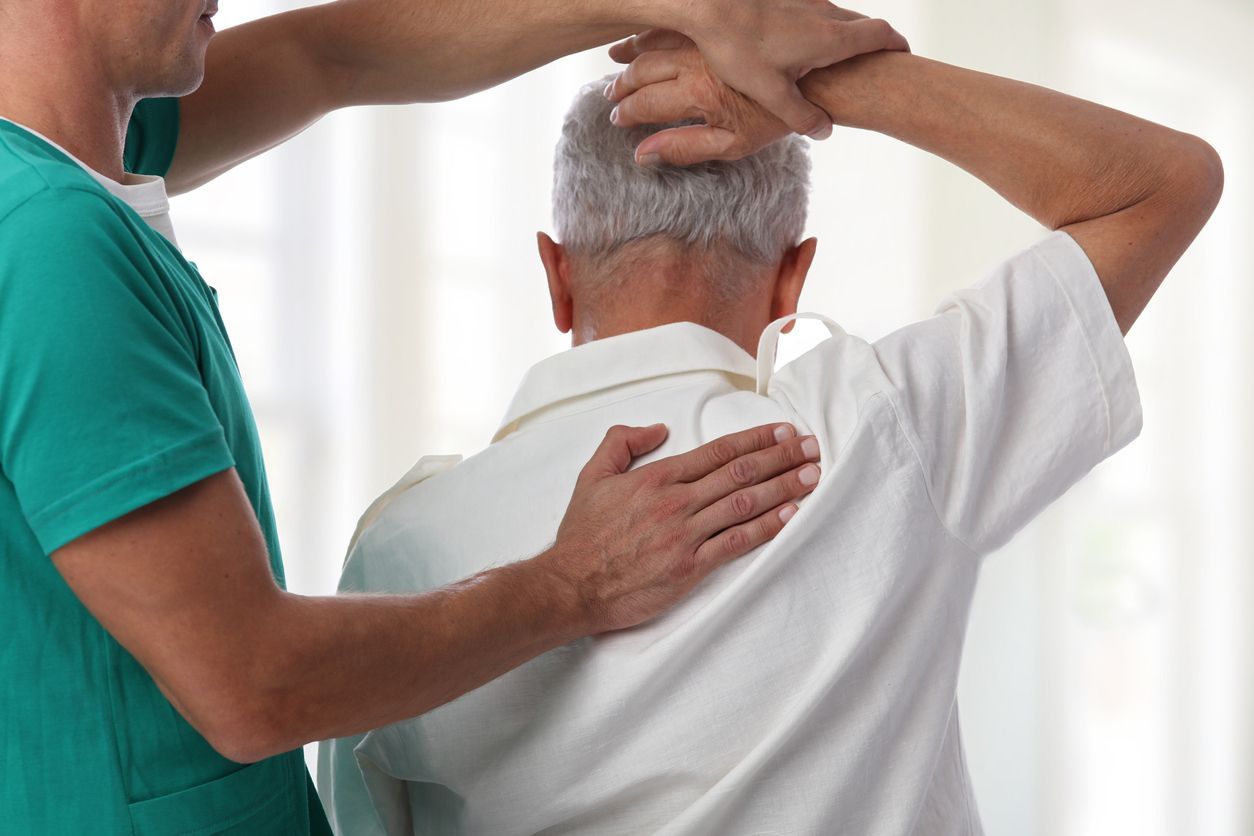 A healthcare professional in green scrubs examines a patient’s shoulder and back movement during a physical therapy session.