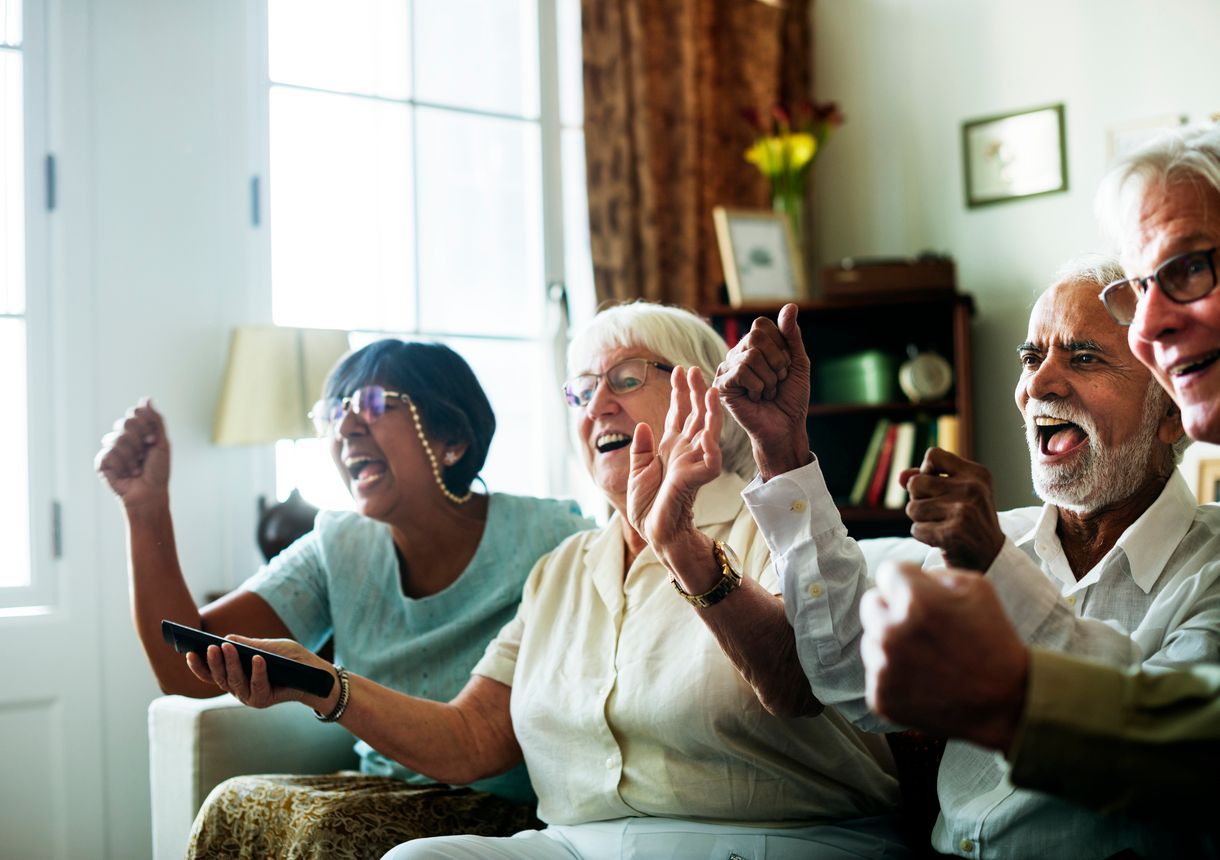 Four people sitting on a couch reacting excitedly while watching television in a brightly lit living room.