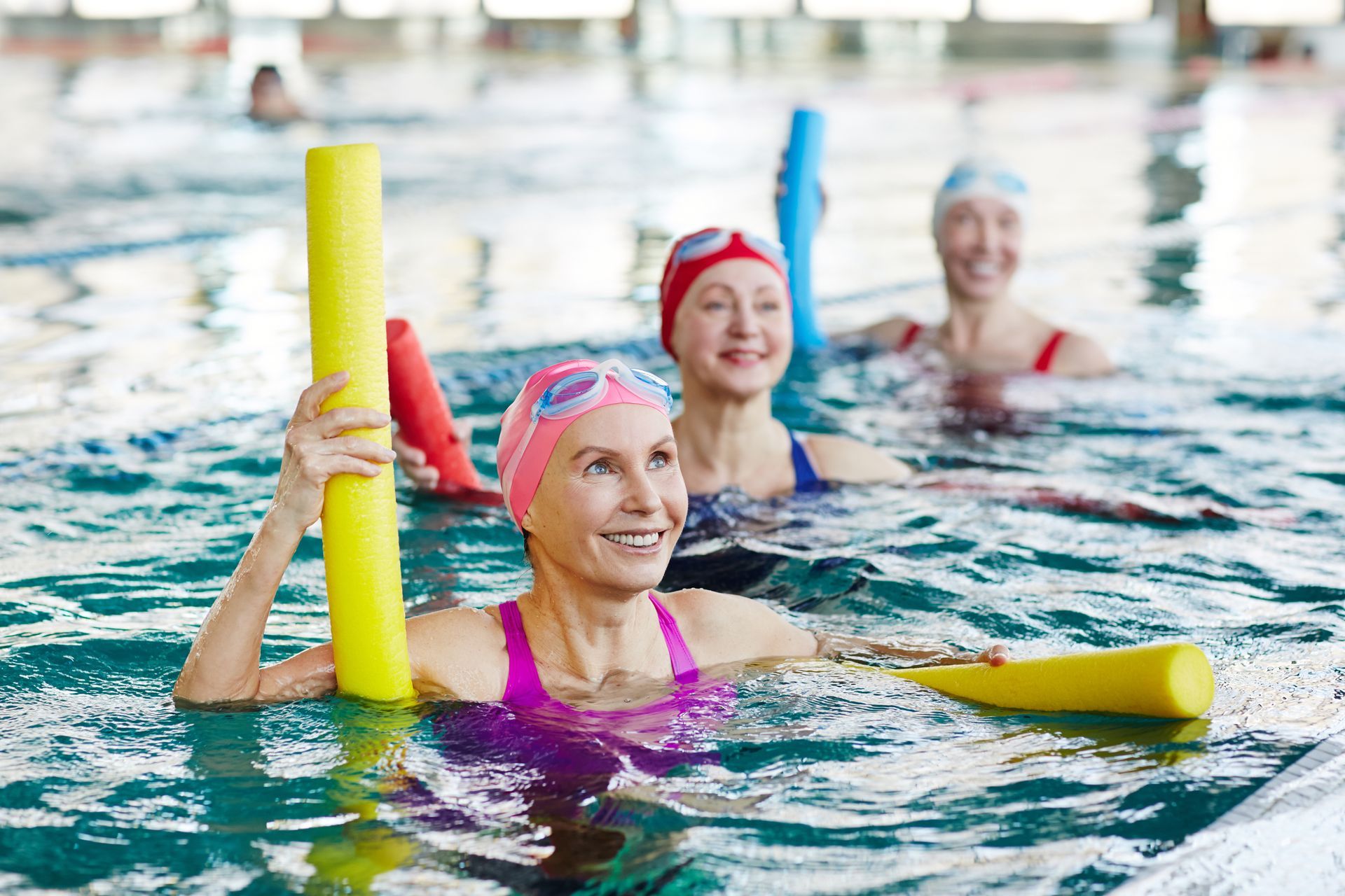 People with swim caps and goggles participate in a water aerobics class in a pool using colorful foam noodles.