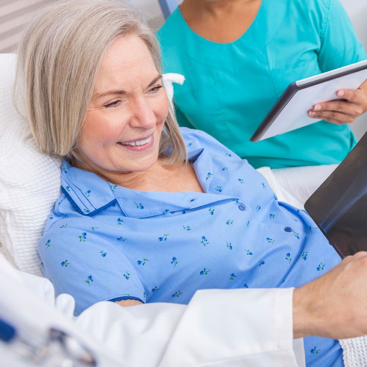 A patient in a hospital bed smiling while a medical professional in scrubs holds a tablet nearby.