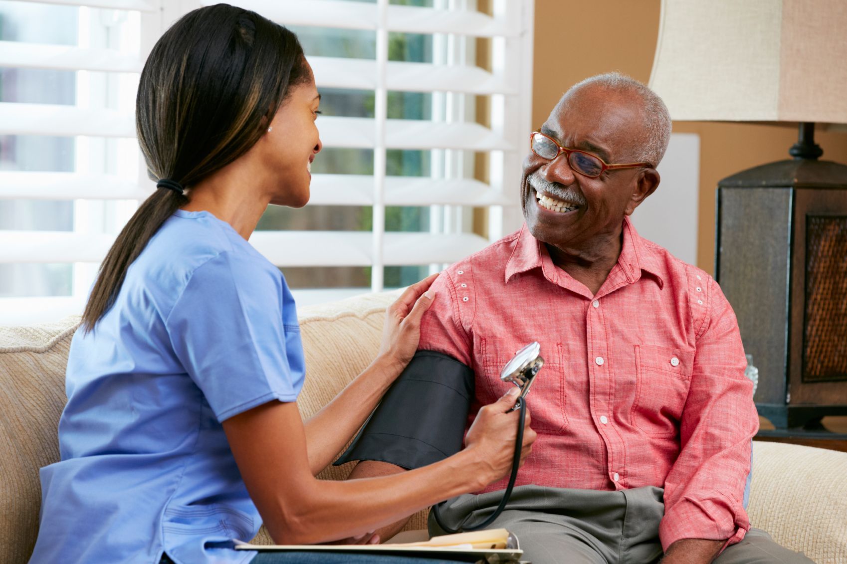 A healthcare worker in blue scrubs checks a patient's blood pressure while the patient smiles in a bright, homey room.