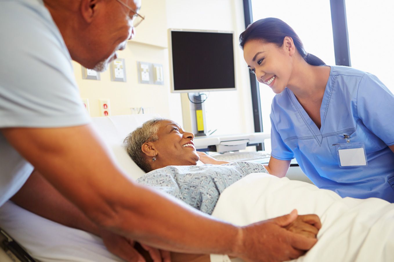 A healthcare professional in blue scrubs speaks kindly to a patient in a hospital bed while a visitor holds their hand.
