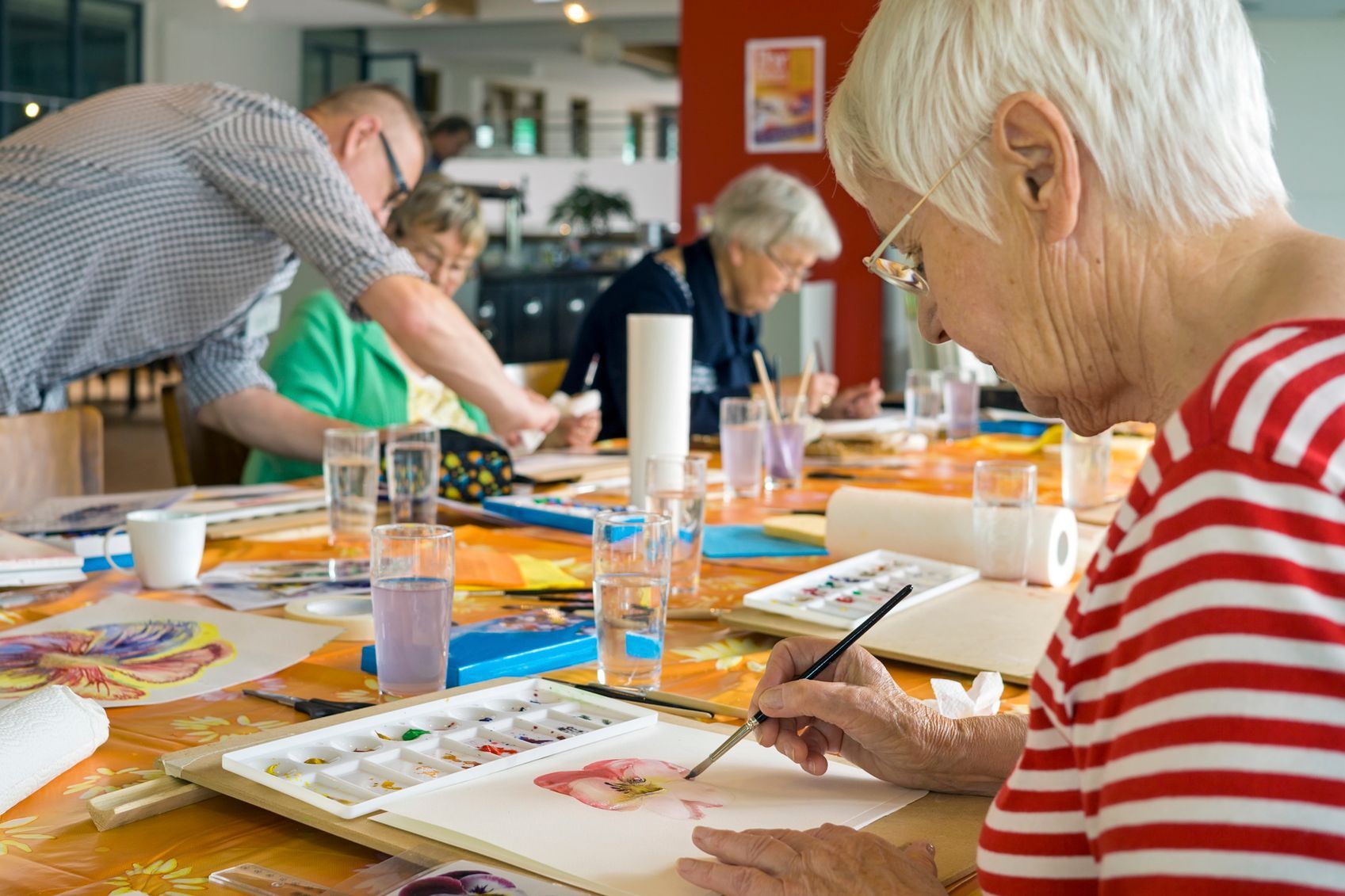 People in a bright room painting with watercolors at a large table covered with an orange patterned cloth.