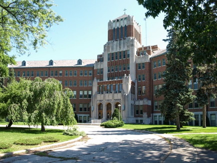 A large, red brick building with an Art Deco central tower stands behind a paved driveway surrounded by trees and lawn.