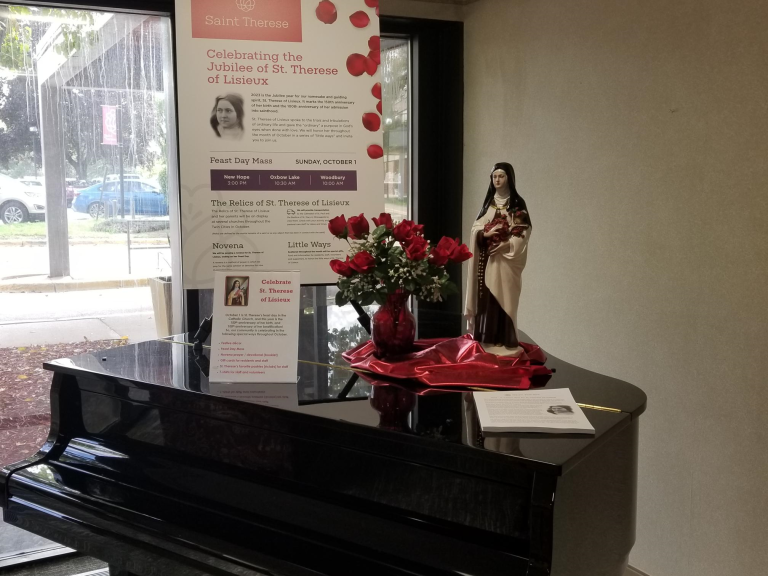 A statue and bouquet of red roses sit atop a grand piano in front of a religious display featuring St. Thérèse of Lisieux.