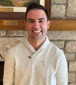A smiling person wearing a beige shawl-collar sweater, posed in front of a stone wall with wooden shelving and a plaque.