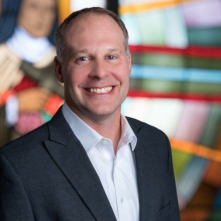 A smiling man in a dark blazer and light button-down shirt stands in front of a colorful, blurred stained-glass window.