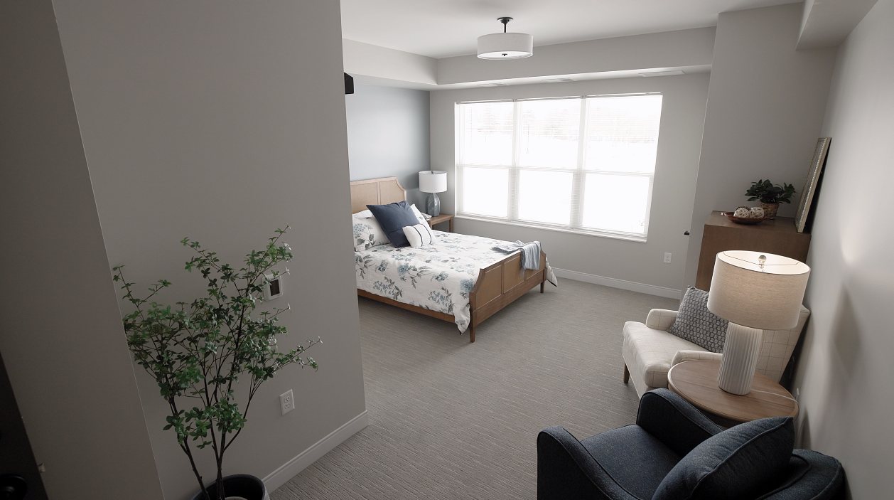 A neutral-toned bedroom with a bed, two armchairs, a side table with a lamp, and a potted plant in the foreground.