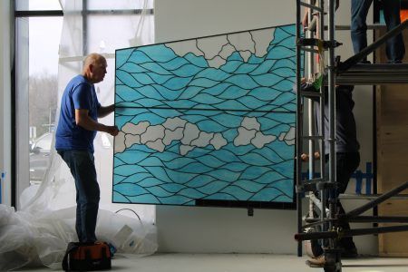 Workers install a large rectangular panel featuring a blue-and-white abstract, wave-like pattern in a bright room.