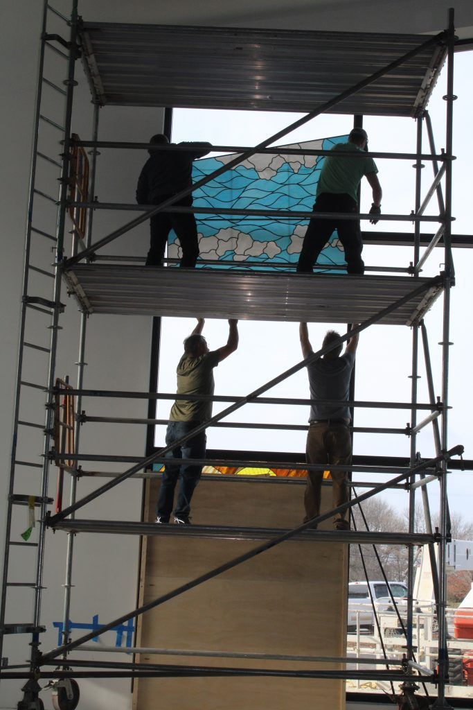 Four people work on a multi-level scaffolding structure, installing a blue and white patterned pane into a window frame.