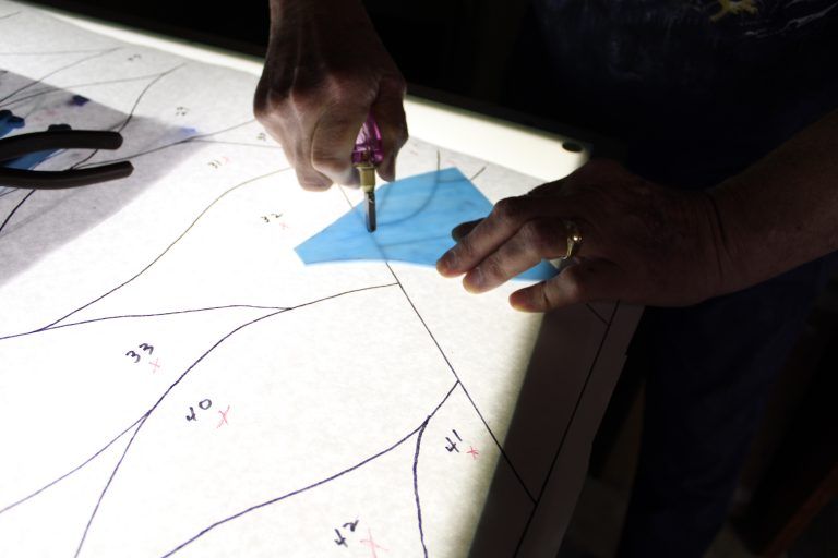Hands use a glass cutter on a blue glass piece placed over a numbered pattern on a light table.
