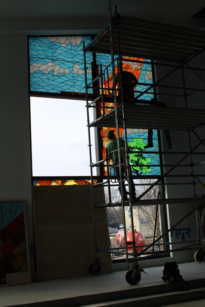 Workers on scaffolding install a stained-glass window panel featuring blue sky, orange flames, and green trees.