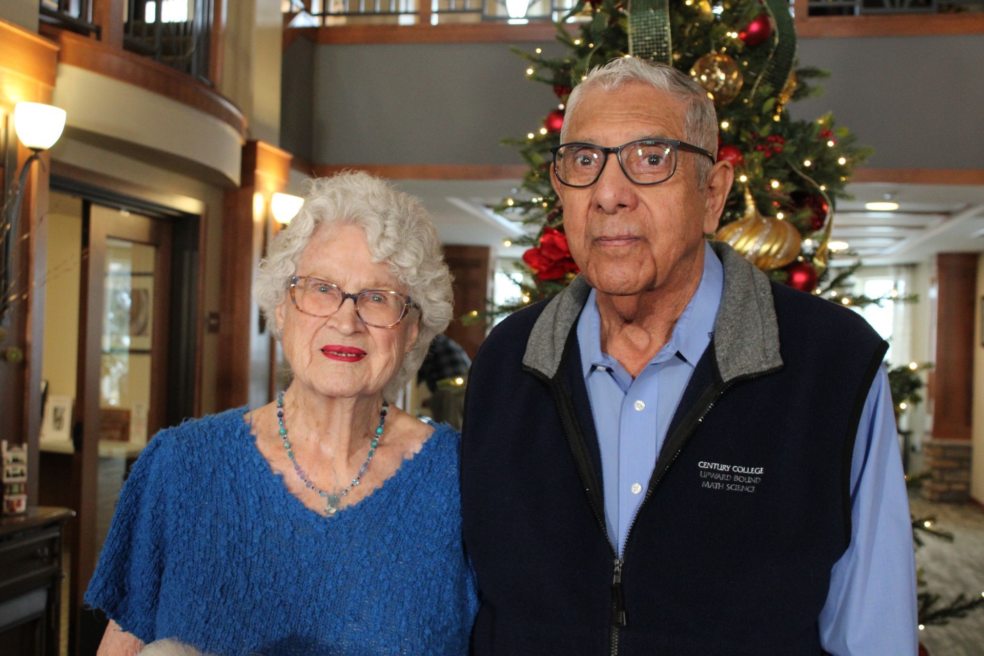 Two people standing indoors in front of a decorated Christmas tree.